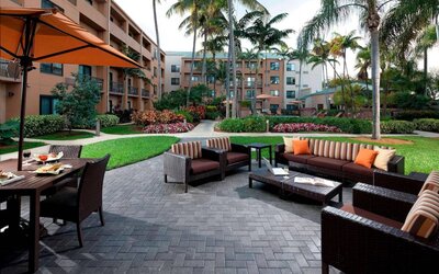 Patio area furnished with sofa and armchair seating and shaded table and chairs, close to pleasant greenery at Sonesta Select Miami Lakes.