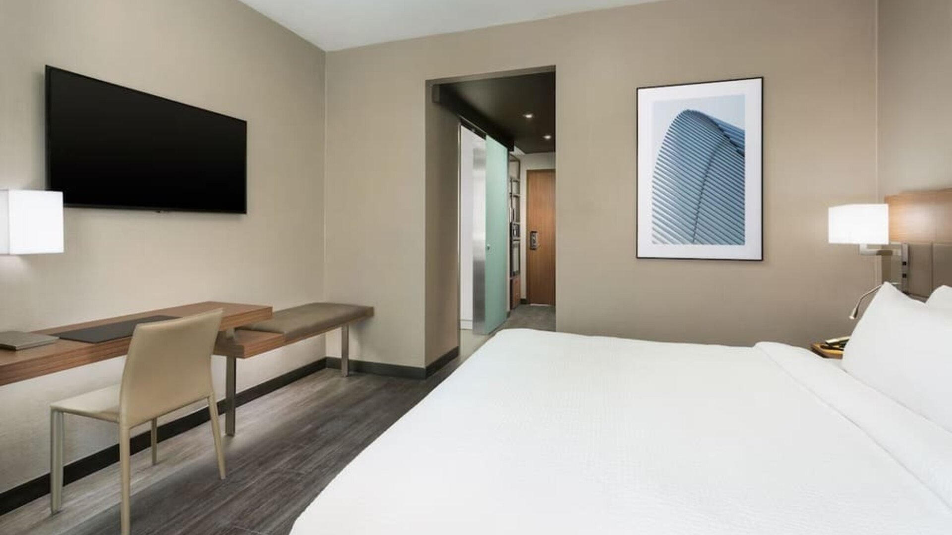 Modern guest room featuring a plush king bed, dedicated workspace, wall-mounted TV, contemporary furnishings, and a bright, minimalist design.