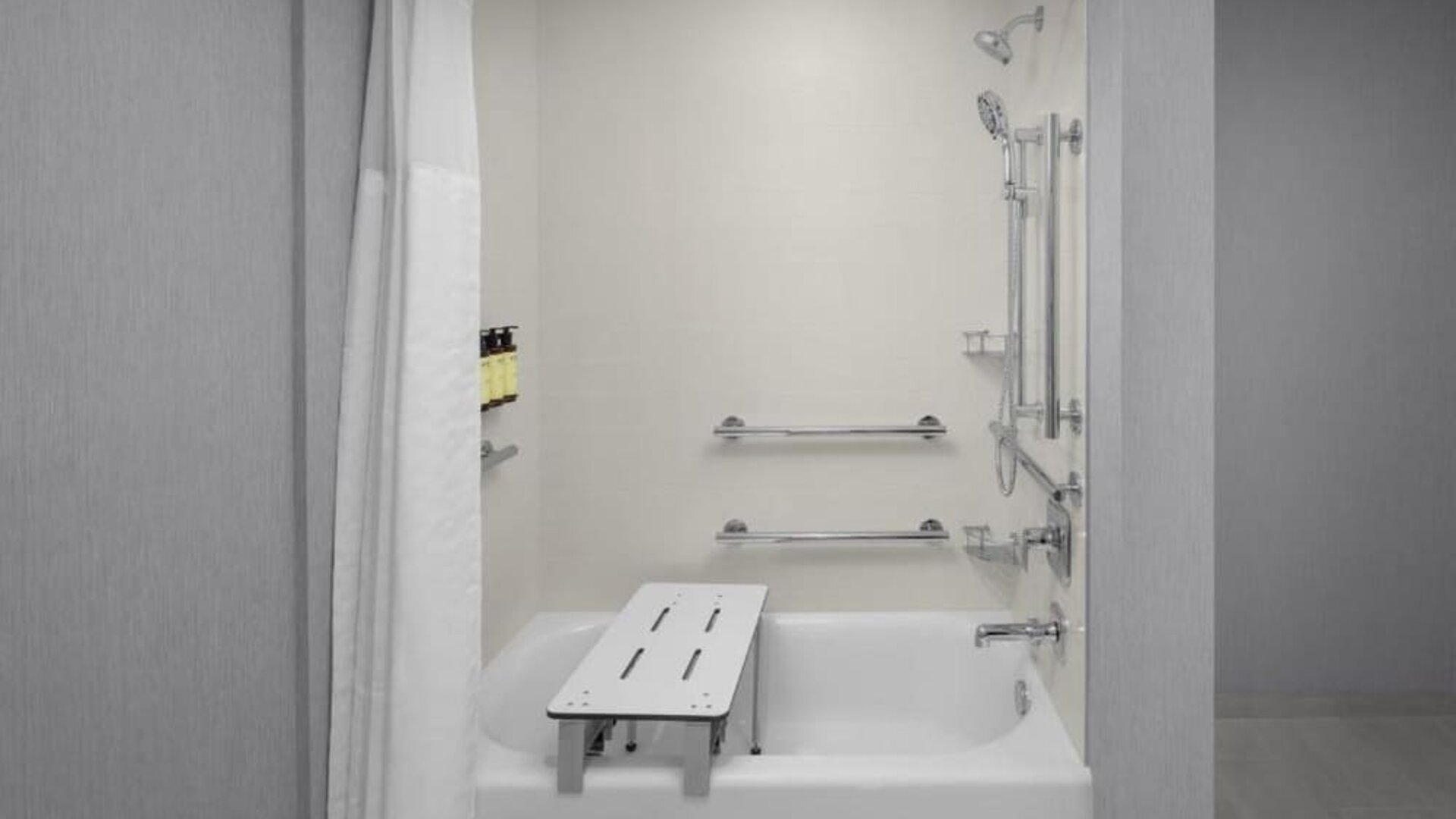 Accessible bathroom with bathtub, grab bars, handheld showerhead, and transfer bench at Hyatt Place Bozeman Yellowstone Airport.