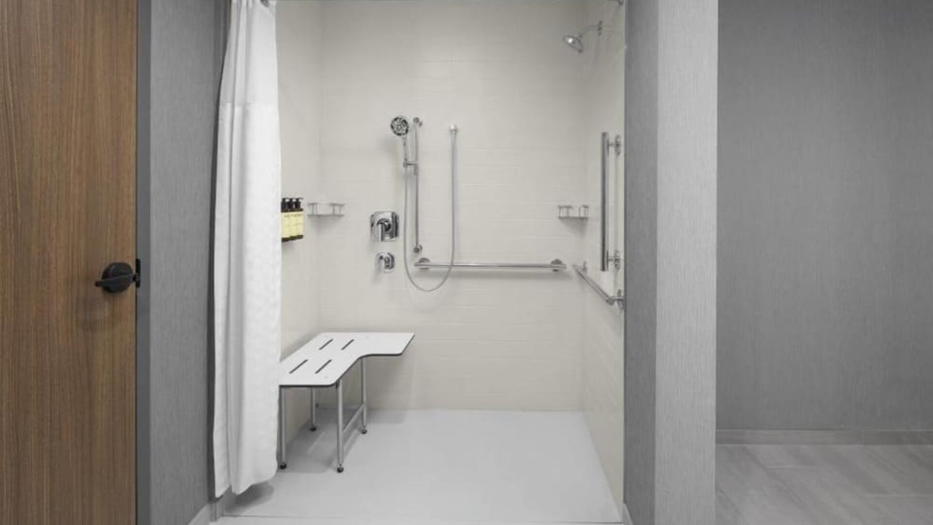Accessible roll-in shower with grab bars, handheld showerhead, and built-in transfer bench at Hyatt Place Bozeman Yellowstone Airport.