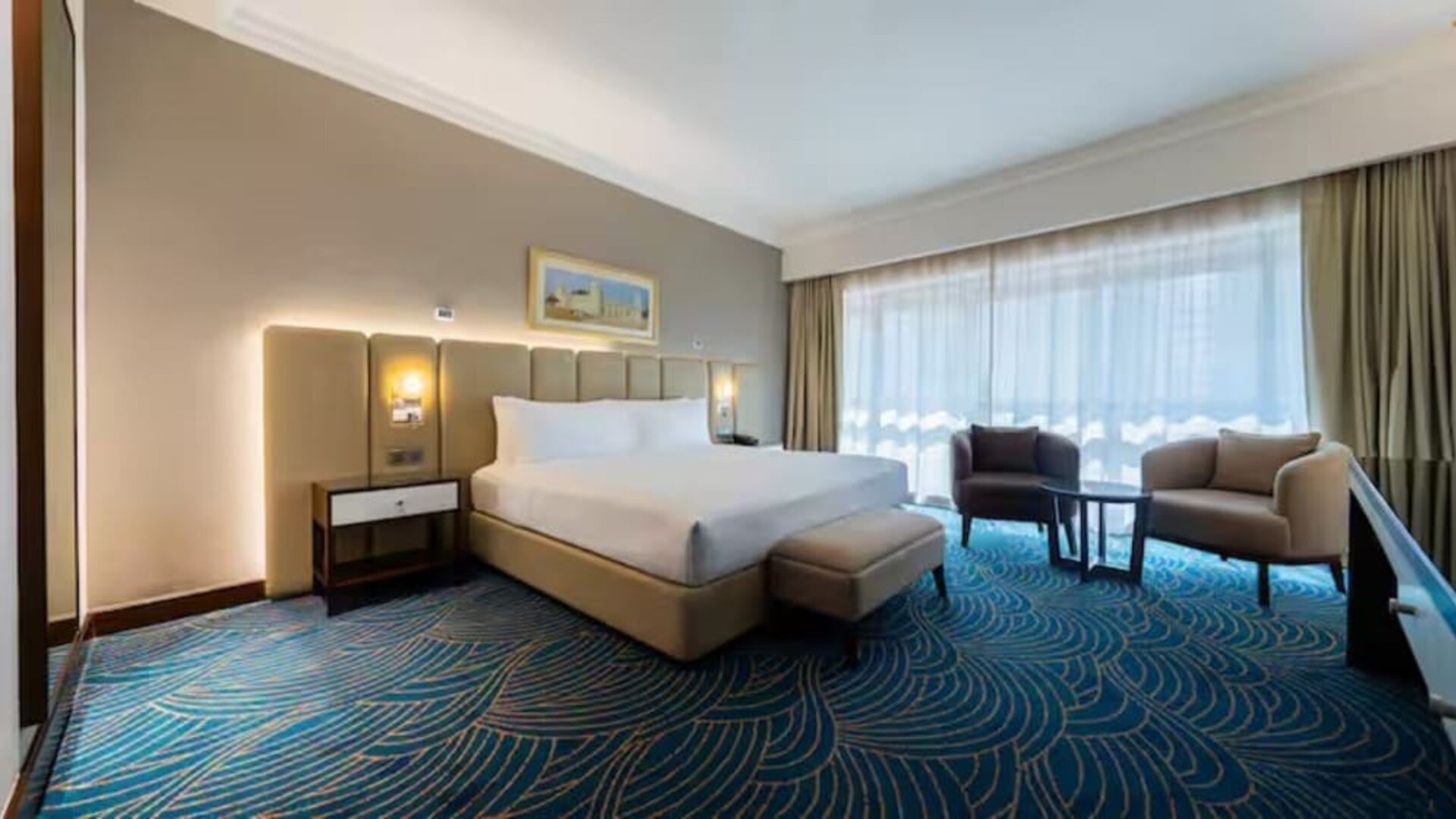 Spacious king room with floor-to-ceiling windows, lounge seating, patterned carpet, and natural daylight.