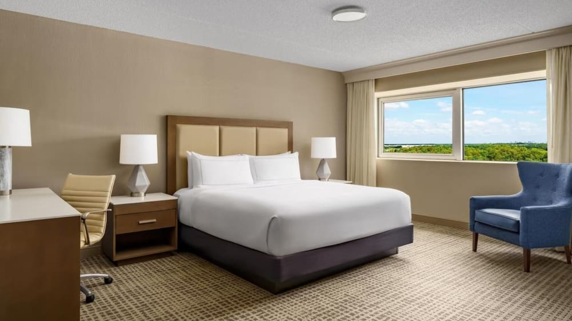 Guest room with king bed, desk, armchair, and private balcony overlooking the pool area.