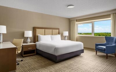 Guest room with king bed, desk, armchair, and private balcony overlooking the pool area.