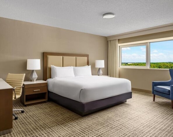 Guest room with king bed, desk, armchair, and private balcony overlooking the pool area.