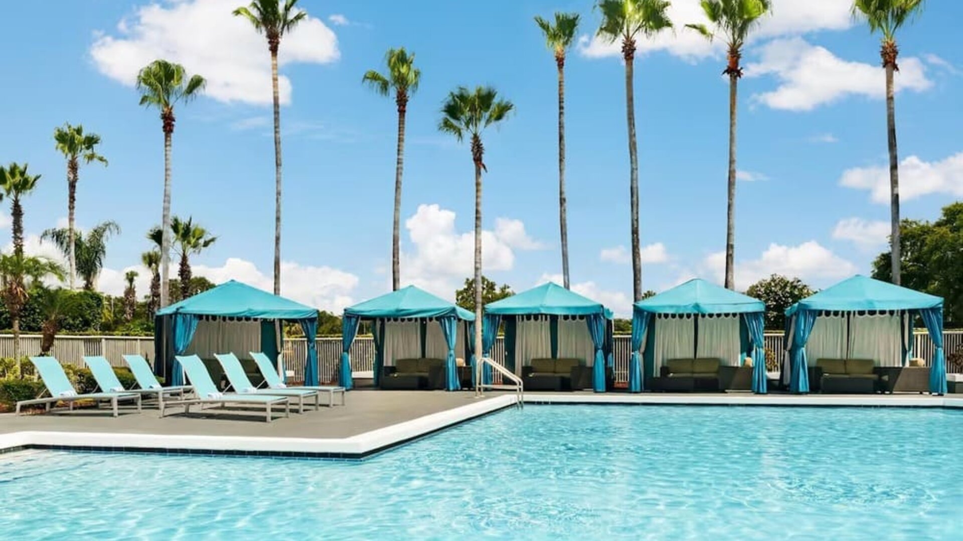 Outdoor swimming pool with turquoise cabanas, lounge chairs, palm trees, and sunny resort atmosphere.