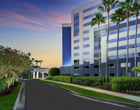Palm-lined hotel entrance with landscaped gardens, curved driveway, and multi-story exterior at sunset.