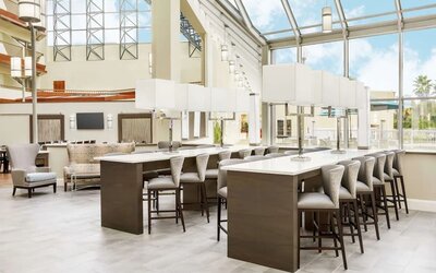Bright atrium workspace with high-top communal tables, cushioned seating, floor-to-ceiling windows, and abundant natural light.