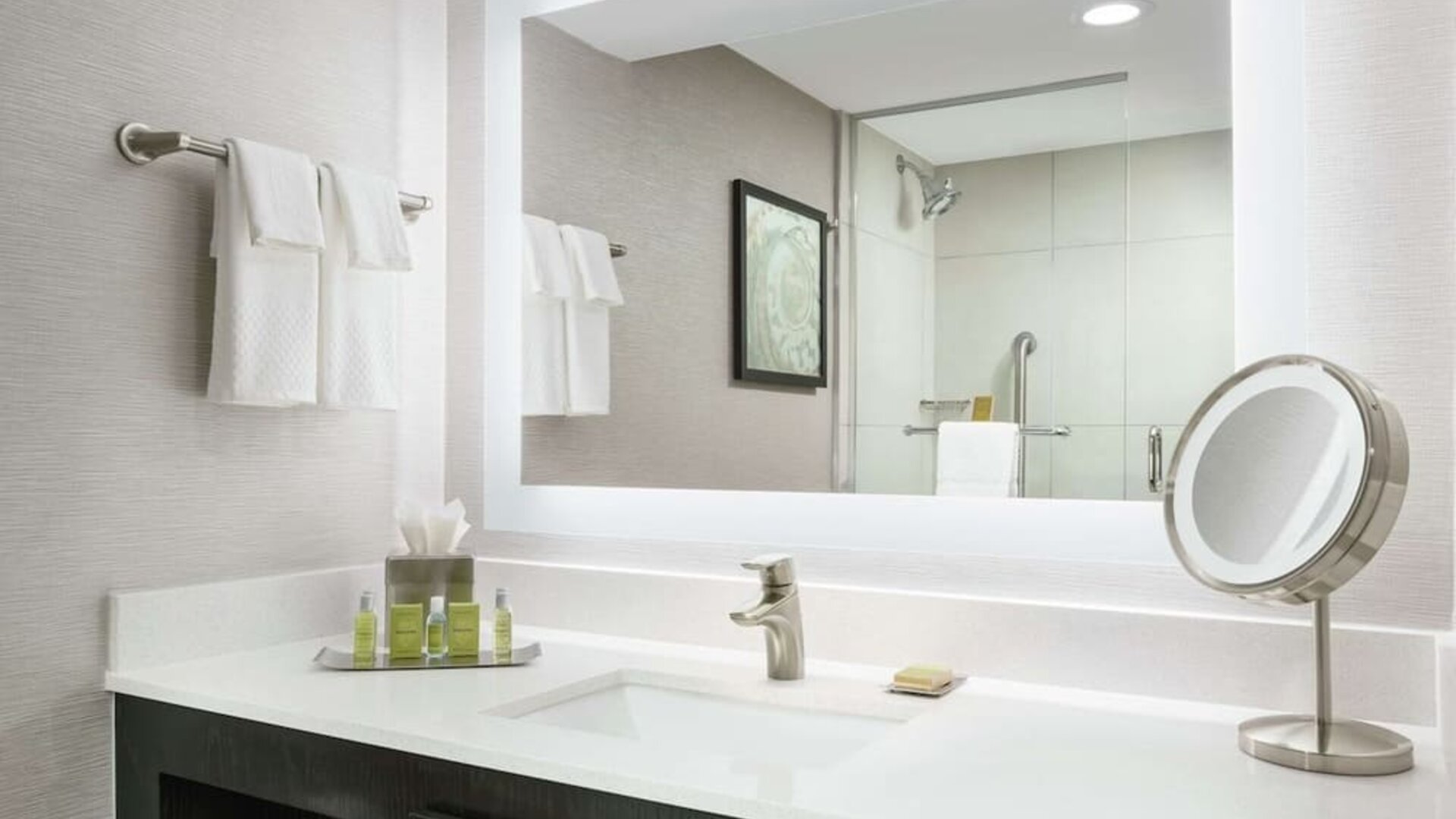 Contemporary bathroom vanity with large mirror, bright lighting, toiletries, towels, and glass-enclosed shower.