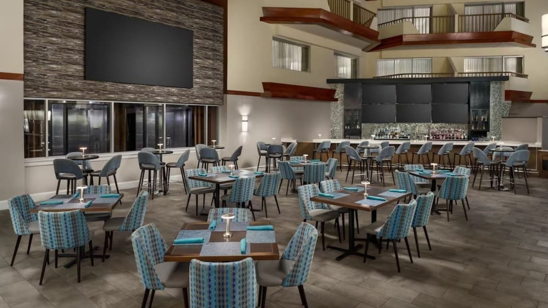 Spacious hotel restaurant with dining tables, upholstered chairs, atrium-style architecture, and adjacent bar area.