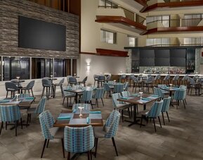 Spacious hotel restaurant with dining tables, upholstered chairs, atrium-style architecture, and adjacent bar area.