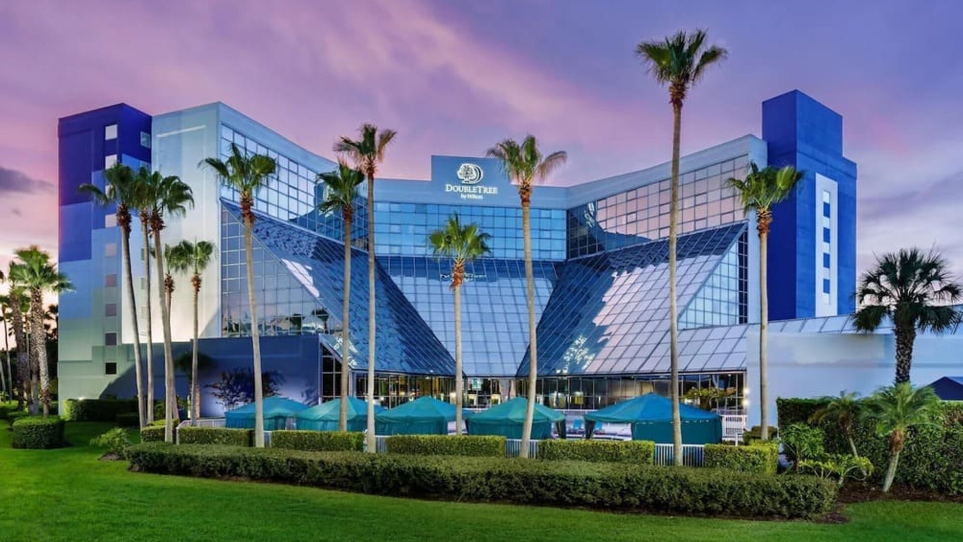 Glass-fronted hotel exterior surrounded by palm trees and manicured lawns under a colorful evening sky.