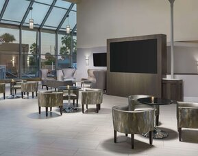 Elegant lobby lounge with armchairs, cocktail tables, soaring glass ceiling, and large display screen.