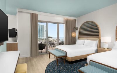 Guest room with two beds, patterned rug, balcony access, and views toward the beach and nearby buildings.