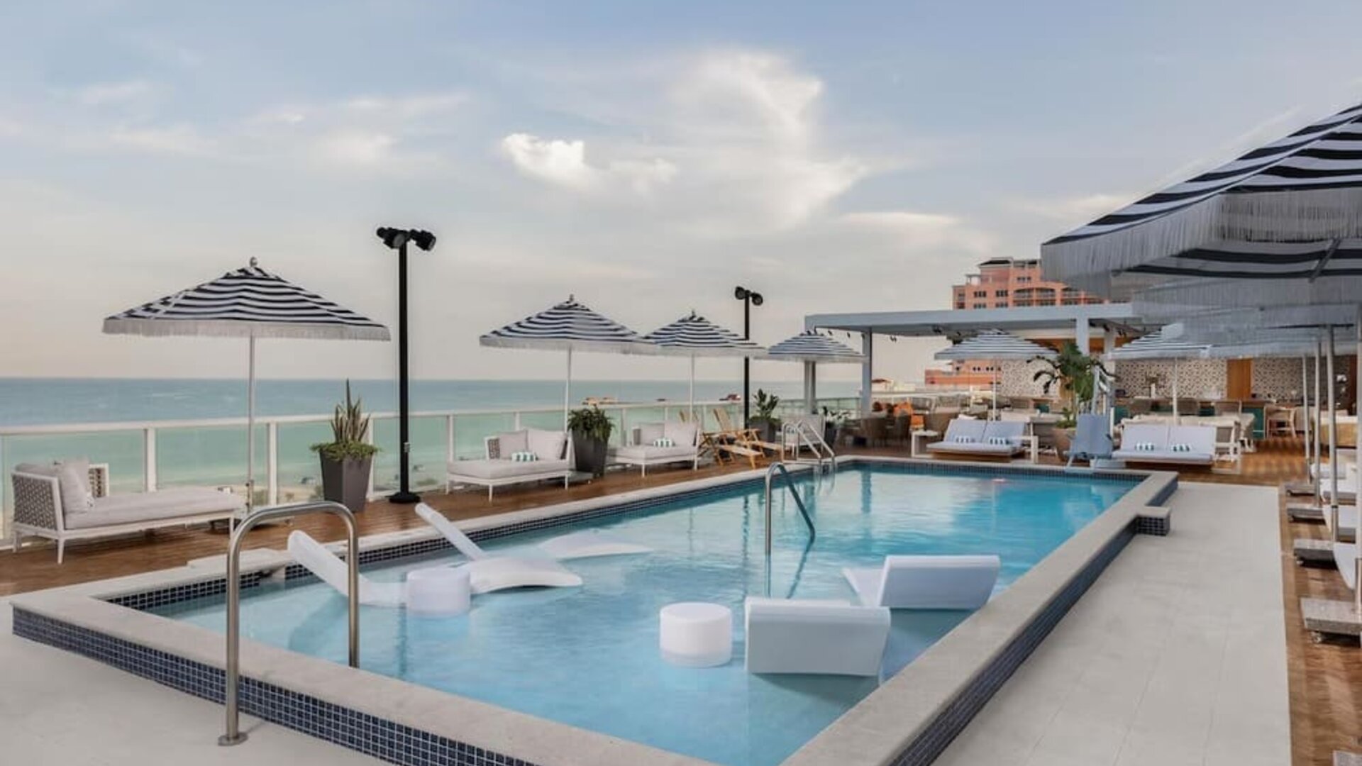Poolside loungers beneath striped umbrellas overlooking the ocean during a soft coastal sunset.