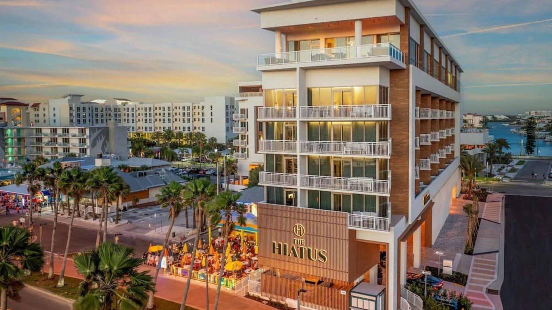 Modern hotel exterior at sunset featuring balconies, illuminated façade, palm-lined streets, and outdoor dining below.