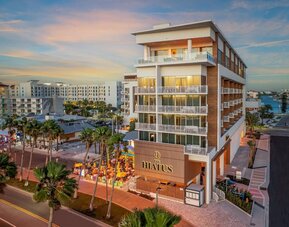 Modern hotel exterior at sunset featuring balconies, illuminated façade, palm-lined streets, and outdoor dining below.