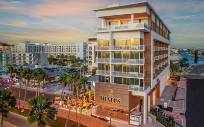 Modern hotel exterior at sunset featuring balconies, illuminated façade, palm-lined streets, and outdoor dining below.