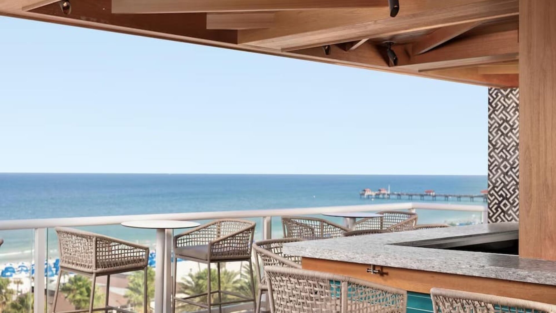 Open-air rooftop bar with woven stools, ocean views, beach below, and distant pier extending into the water. 