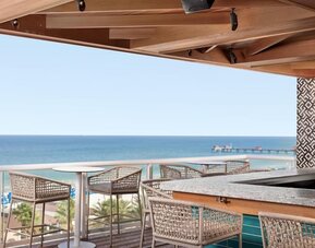 Open-air rooftop bar with woven stools, ocean views, beach below, and distant pier extending into the water. 