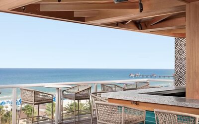 Open-air rooftop bar with woven stools, ocean views, beach below, and distant pier extending into the water. 