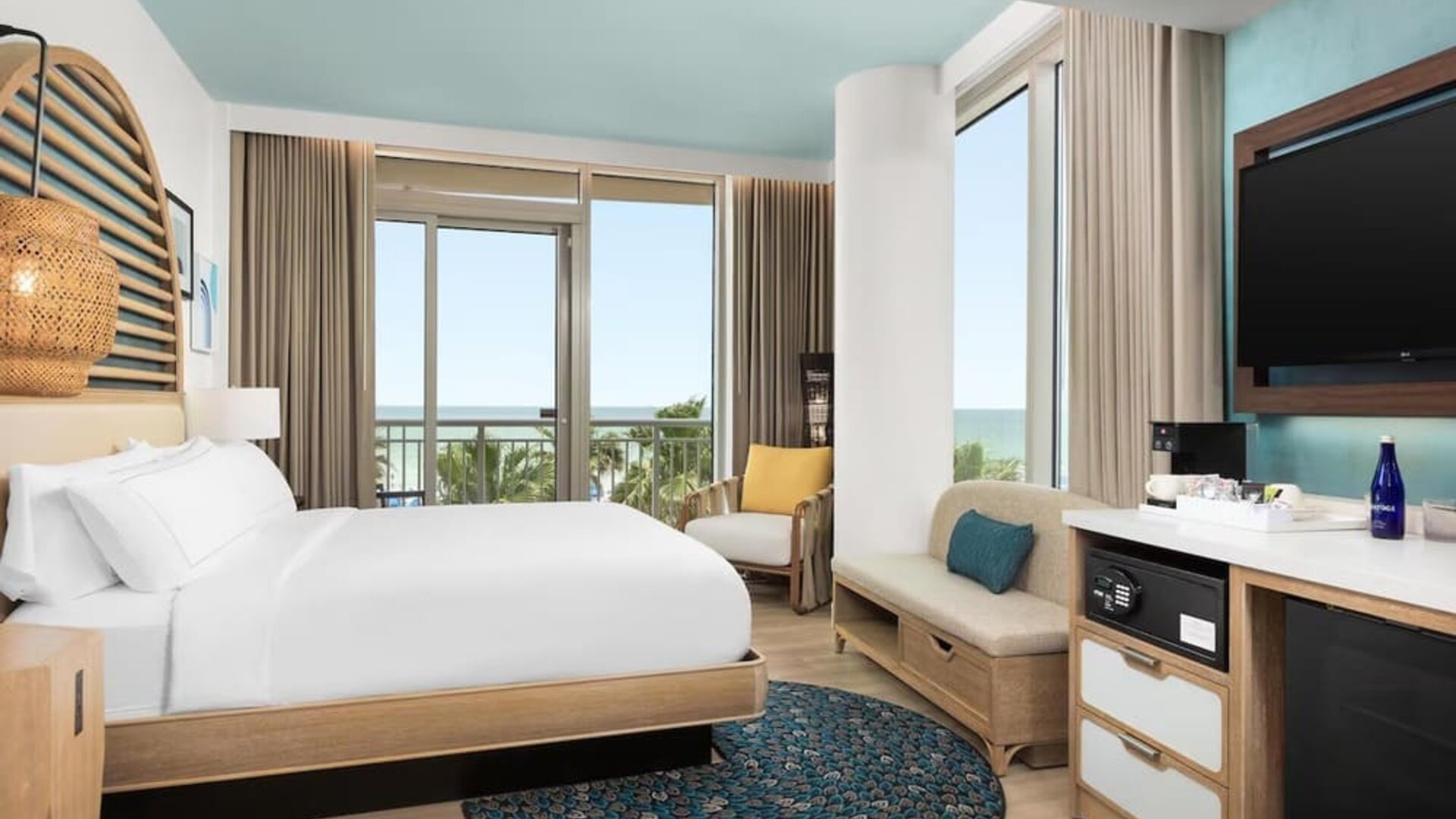 Contemporary guest room with king bed, seating area, large TV, and balcony offering partial ocean views.