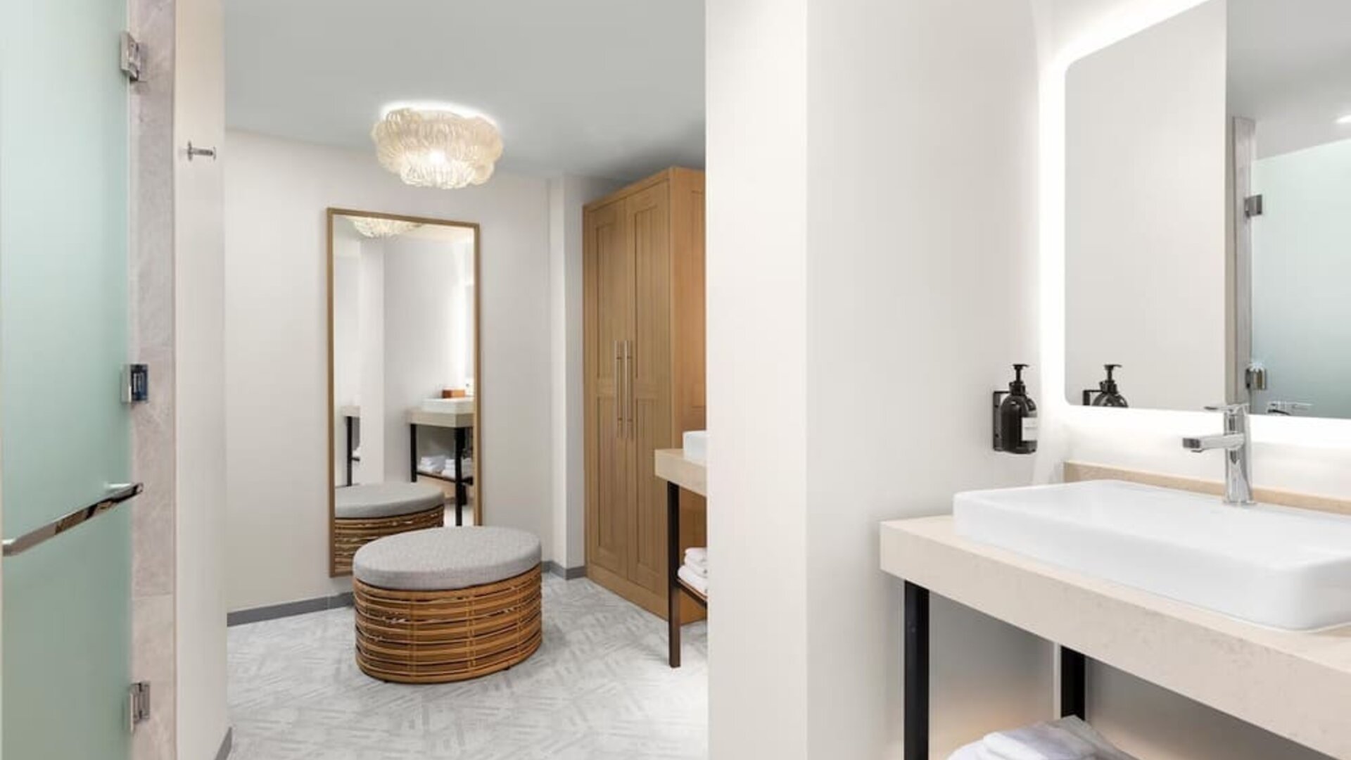Spacious bathroom and dressing area with illuminated mirror, vessel sink, woven ottoman, and wood wardrobe.