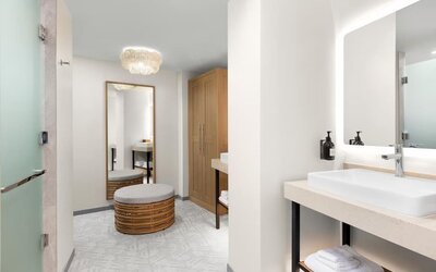 Spacious bathroom and dressing area with illuminated mirror, vessel sink, woven ottoman, and wood wardrobe.