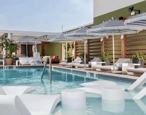 Rooftop pool deck with in-water loungers, striped umbrellas, ocean views, and comfortable relaxation areas.