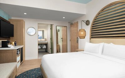Bright guest room with king bed, wood accents, wall-mounted TV, and open vanity area leading to bathroom.