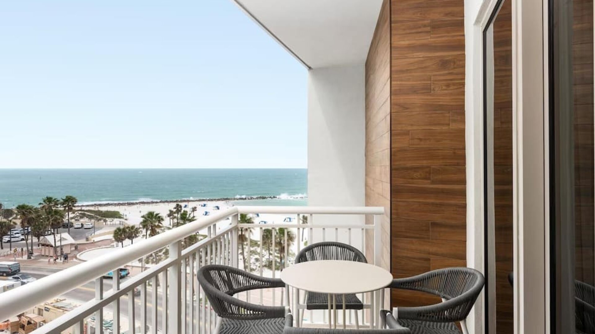 Balcony with round table and four woven chairs overlooking white-sand beach, palm trees, and turquoise ocean.