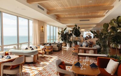 Upscale restaurant and lounge with ocean-facing windows, stylish seating, lush plants, and natural daylight.