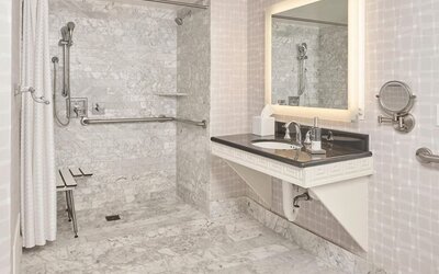 Accessible marble bathroom with roll-in shower, grab bars, illuminated vanity, and spacious design at Hyatt Centric Delfina Santa Monica.
