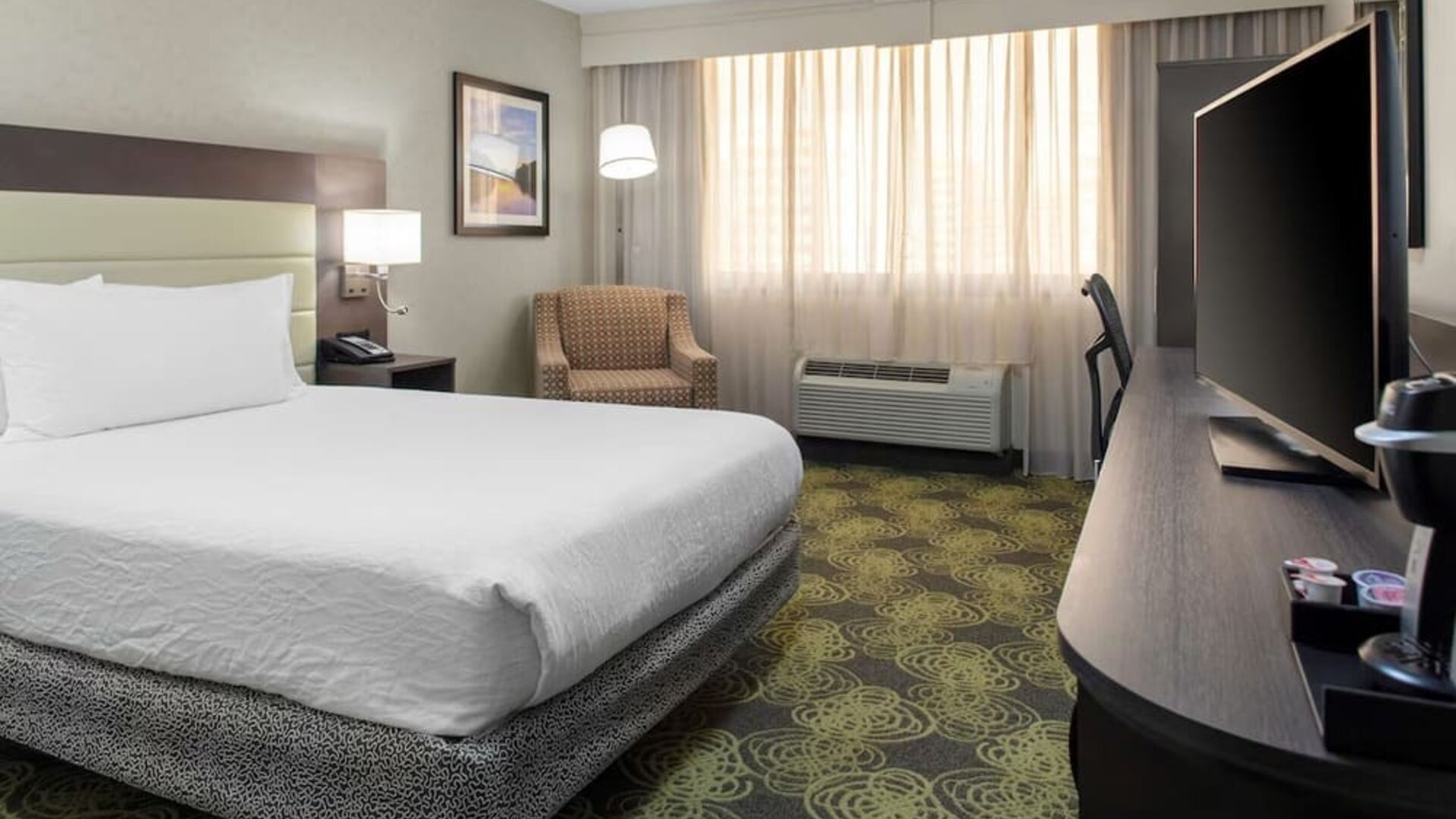 King guest room with plush bed, HDTV, armchair, workspace, and large windows providing natural daylight.