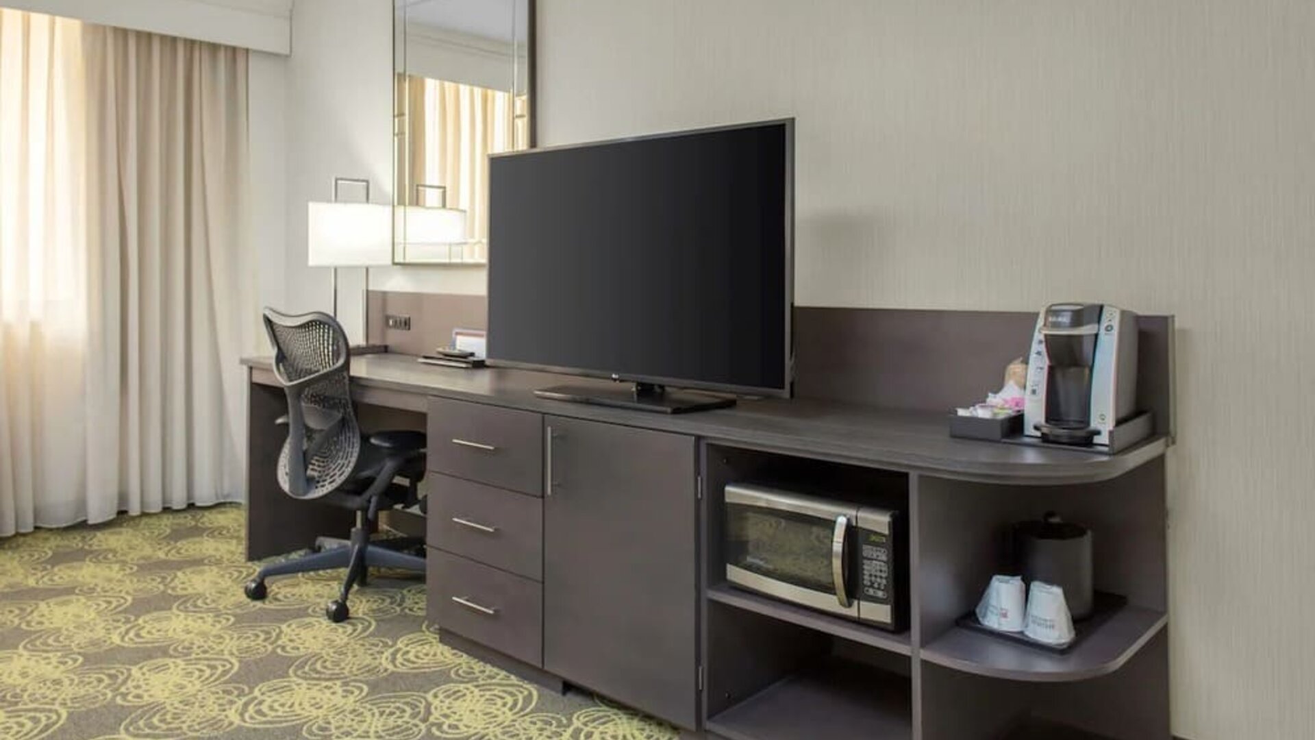 Guest room featuring a large HDTV, ergonomic desk, coffee maker, microwave, and workspace for business travelers.