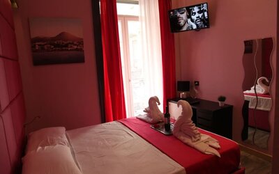 Double room with red curtains, balcony access, towel swans and wall-mounted TV at Dolce Vita Rooms & Breakfast.