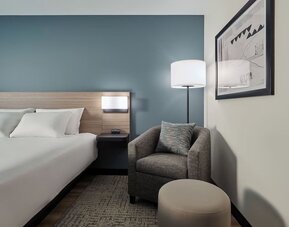 Cozy guest room corner with a plush armchair, ottoman, floor lamp and comfortable king bed at Hyatt Select Philadelphia Airport.
