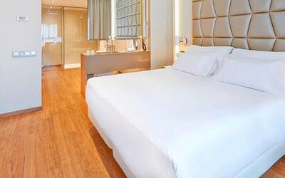 Contemporary guest room featuring a large bed, geometric upholstered headboard, wood flooring and open bathroom area at NH Collection Barcelona Pódium.