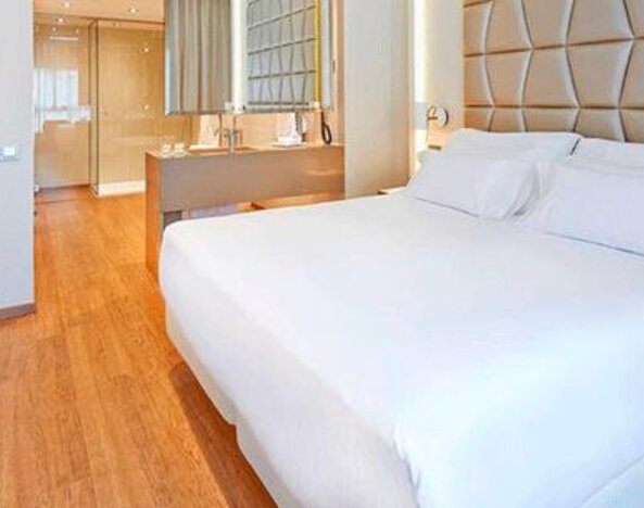 Contemporary guest room featuring a large bed, geometric upholstered headboard, wood flooring and open bathroom area at NH Collection Barcelona Pódium.