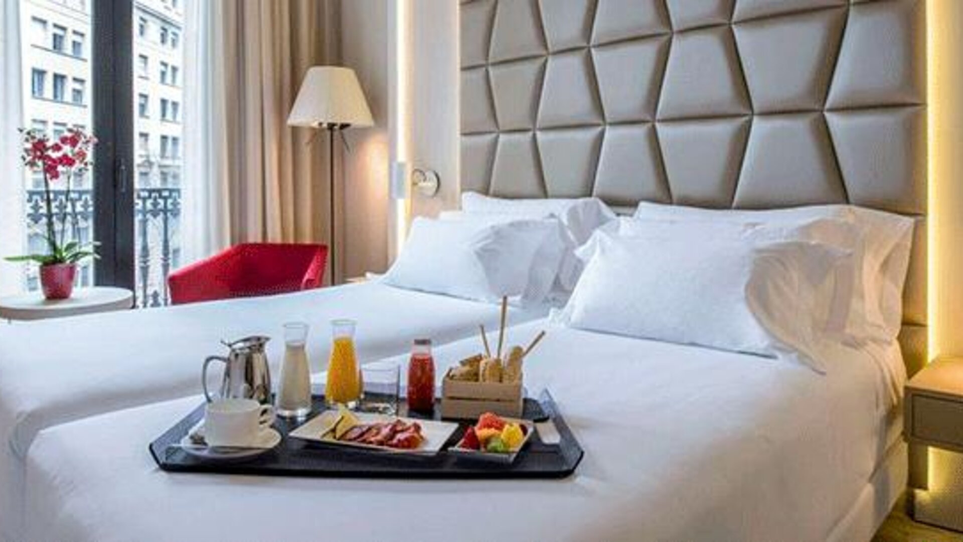 Guest room featuring a large bed, breakfast tray, red accent chair and natural light from the balcony doors at NH Collection Barcelona Pódium.