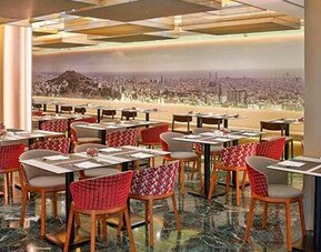 Spacious restaurant featuring dining tables, red patterned chairs and a panoramic city mural at NH Collection Barcelona Pódium.