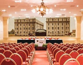 Large conference hall with rows of red chairs, a presentation stage, chandeliers, and a decorative mural backdrop at Hotel Geneve.