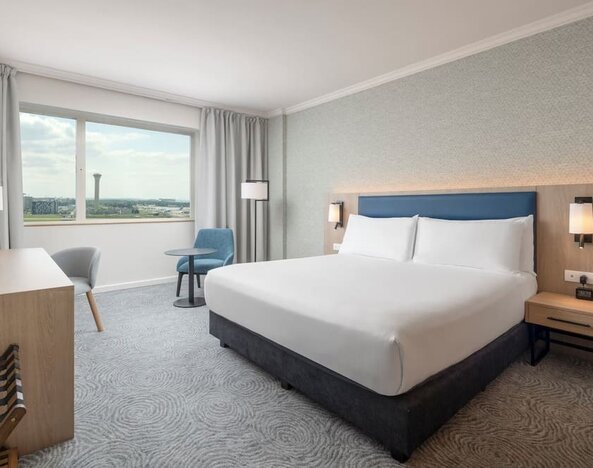 Modern king guest room with bed, large television, long work desk, armchairs, side table and floor-to-ceiling curtains.