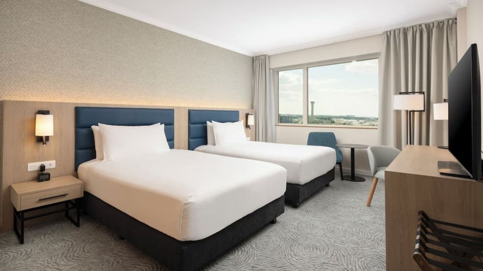 Bright twin guest room with two beds, airport-view window, desk, television, chairs and contemporary furnishings.