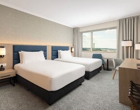 Bright twin guest room with two beds, airport-view window, desk, television, chairs and contemporary furnishings.