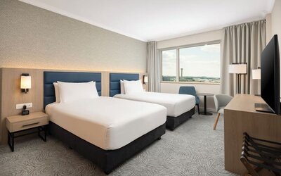 Bright twin guest room with two beds, airport-view window, desk, television, chairs and contemporary furnishings.