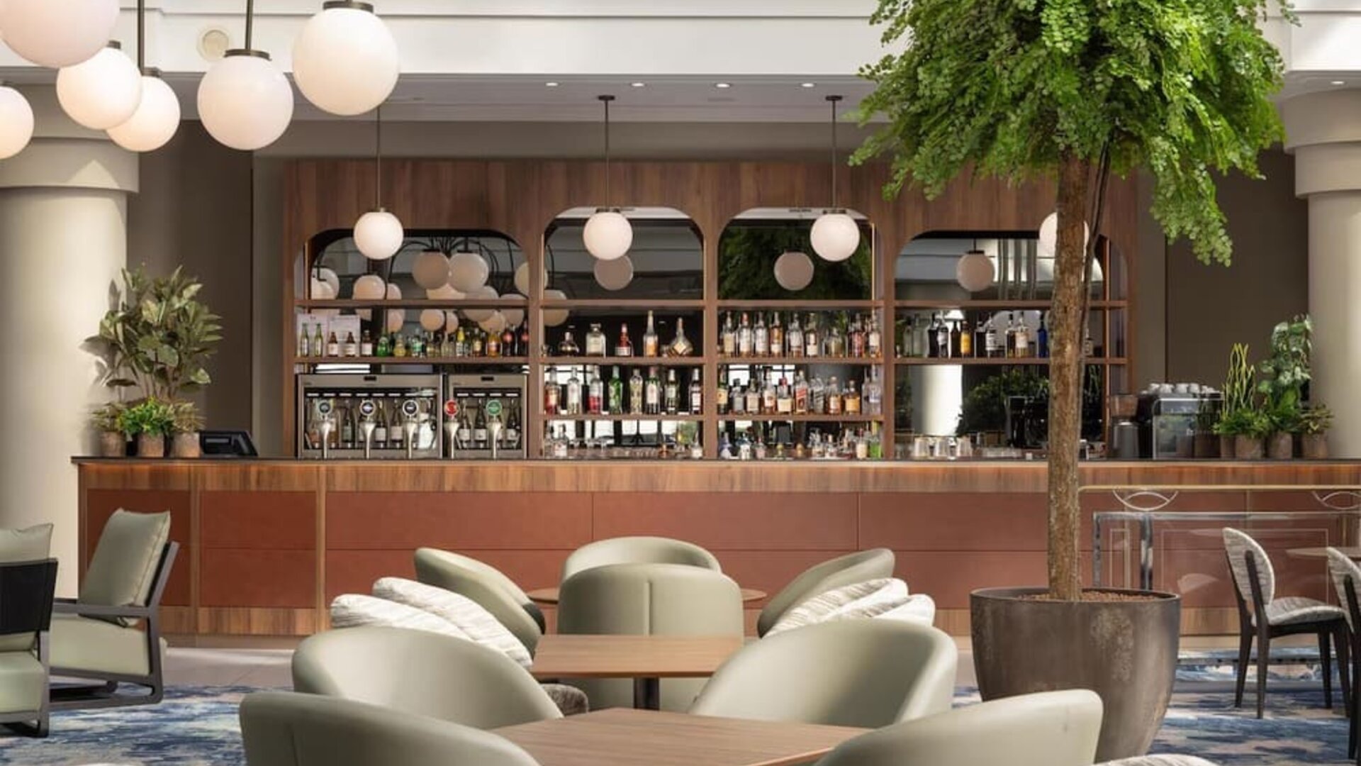 Contemporary hotel bar with upholstered lounge seating, wooden tables, globe pendant lights, plants and stocked bar.