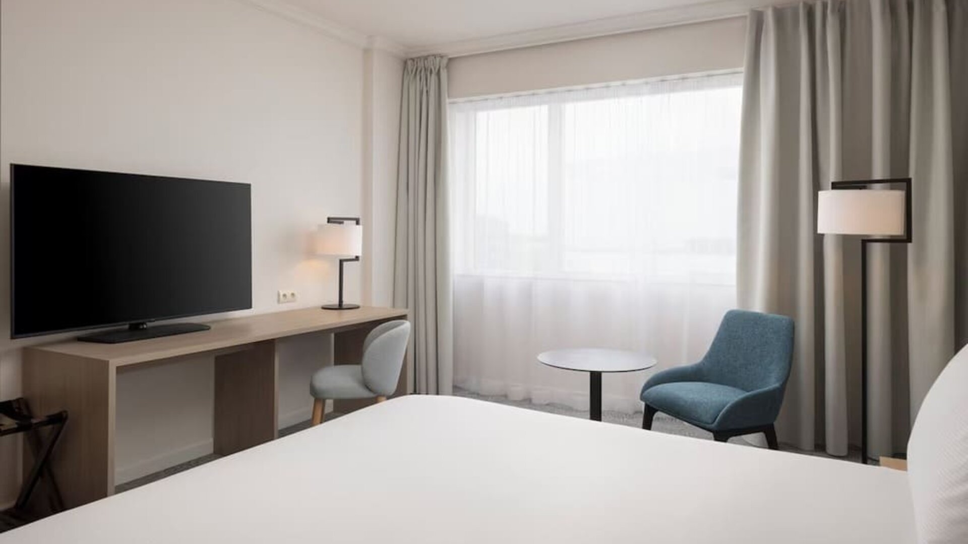 Modern king guest room with bed, large television, long work desk, armchairs, side table and floor-to-ceiling curtains.