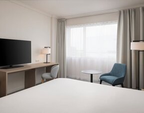 Modern king guest room with bed, large television, long work desk, armchairs, side table and floor-to-ceiling curtains.