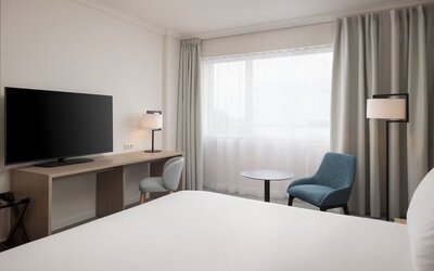 Modern king guest room with bed, large television, long work desk, armchairs, side table and floor-to-ceiling curtains.