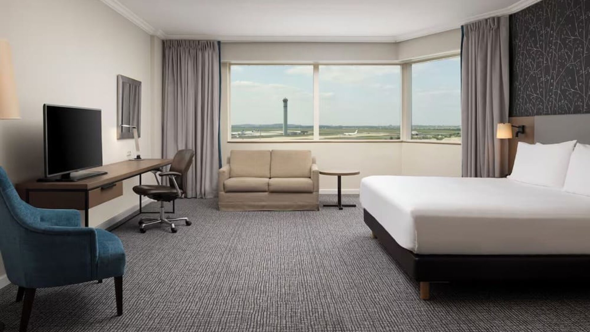 Spacious hotel room with king bed, sofa, work desk, television, armchair and panoramic windows overlooking the airport.